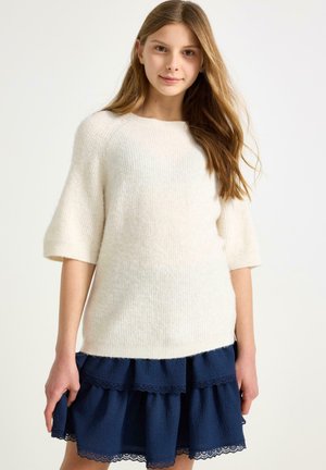 Świetlisty kremowy dziany sweter z krótkimi rękawami, połączony z warstwową granatową spódnicą z falbankami i koronkowymi akcentami.