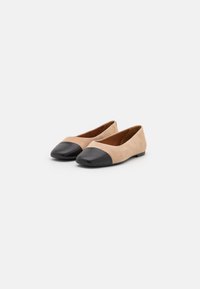 Beige suede flats with black leather toe caps. Smooth texture with a rounded shape and a low-profile design. Inside features brown lining.