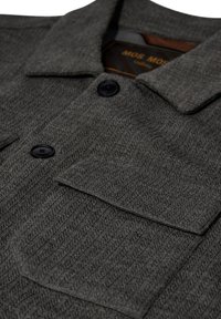 Chaqueta gris texturizada con cuello, dos bolsillos frontales y cierres de botón negros. Etiqueta interior visible con el nombre de la marca.
