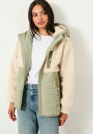 Woman wearing a green quilted and cream sherpa fleece jacket over a white shirt and blue jeans, standing against a plain white wall.