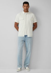 Korte mouwen, off-white shirt met twee borstzakken over een lichtgrijze t-shirt, gecombineerd met lichtblauwe rechte jeans en grijze sneakers.