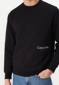 Black cotton sweatshirt with crew neck and long sleeves. Features white "Calvin Klein" logo on the front. Soft texture, casual fit.