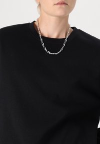 Collar de cadena de plata con eslabones entrelazados, usado sobre un sudadera negra de cuello redondo. Acabado metálico suave que refleja la luz.