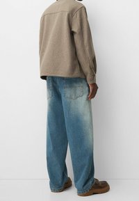 Braune, wollähnliche Außenjacke, lockere Passform. Hellblaue, ausgewaschene, weit geschnittene Jeans mit großen Gesäßtaschen. Braune, klobige Schuhe.