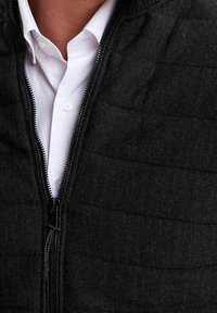 Mężczyzna w białej koszuli zapinanej na guziki pod częściowo zapiętą czarną pikowaną kurtką.