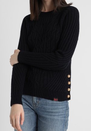 Kvinde iført en sort ribbet sweater med guldknapper i siden og lyseblå jeans, stående med armene over kors.