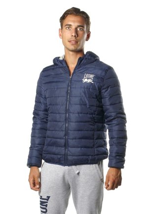 Un homme porte une veste matelassée bleu marine à capuche, avec une fermeture éclair sur le devant et un logo blanc sur la poitrine, assortie à un pantalon de jogging gris, sur fond blanc.