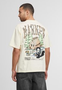 Cremefarvet bomulds T-shirt med et farverigt tegneserieprint af en skunk nær en hytte, med teksten "MARKET" og et citat nedenfor.