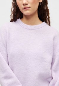 Λιλά πουλόβερ με ριμπ λαιμόκοψη και φαρδιά, απαλά μανίκια. Η υφή φαίνεται πλεκτή, αναδεικνύοντας λεία ύφασμα με ελαφριά λάμψη.