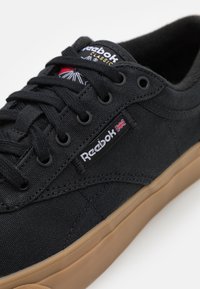 Musta kangaskenkä nauhakiinnityksellä ja kumipohjalla. Varustettu Reebok-logolla ja kontrastivärisillä saumoilla. Sileä pinta.