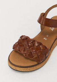 Sandalo in pelle marrone con design della fascia intrecciato, suola in sughero, punta aperta e cinghia alla caviglia marrone per un supporto aggiuntivo.