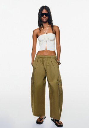 Haut cropped blanc à encolure halter avec un centre froncé, associé à un pantalon cargo large vert olive avec poches latérales. Tongs noires.