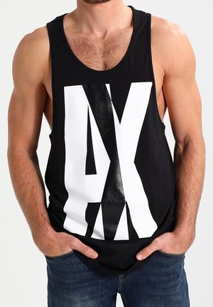 Mand iført en sort tanktop med store hvide og sorte overlappende bogstaver "AX", hænder i lommerne, parret med blå jeans.