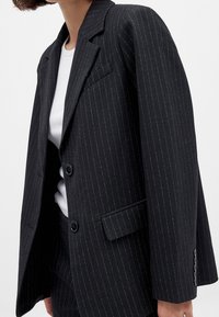 Femme portant un blazer noir à fines rayures avec deux boutons, une poche à rabat et un pantalon assorti sur un top blanc sur fond blanc.