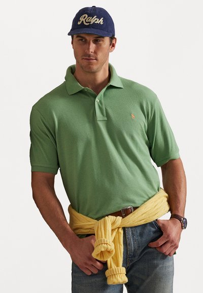 Άνδρας φορώντας πράσινο πόλο μπλουζάκι, μπλε τζιν, κίτρινο πουλόβερ δεμένο στη μέση, μπλε καπέλο με το κείμενο "Ralph" και ρολόι στο χέρι, ποζάροντας με τα χέρια στις τσέπες.