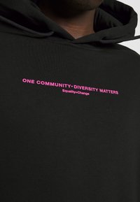 Svart hoodie med rosa text. Texten säger "Ett samhälle - mångfald spelar roll" och "Likhet = förändring." Tillverkad av mjukt tyg, har en huva.