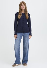 Marineblauwe cardigan met ronde halslijn, knoopsluiting en lange mouwen, gecombineerd met lichtblauwe wijde jeans. Zachte, gebreide textuur.