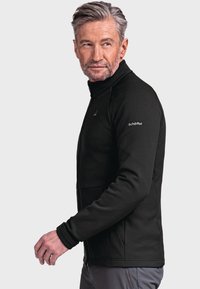 Schwarze Fleecejacke mit hohem Kragen, Reißverschlusstaschen, gerippter Textur und Logodetail am linken Ärmel. Entworfen für Wärme und Komfort.
