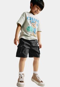 T-shirt à manches courtes de couleur vert clair avec un graphisme de dessin animé et du texte, associé à un short cargo noir et des baskets montantes beiges.