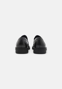Schwarze Lederschuhe mit glatter Oberfläche, runder Spitze und einer niedrigen, strukturierten Gummisohle. Die Rückansicht hebt die minimalen Nähte und das schlanke Design hervor.
