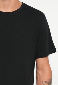 Hombre con una camiseta de cuello redondo de manga corta negra y sencilla, con un tatuaje visible en el brazo derecho inferior, contra un fondo blanco.