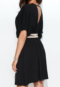 Musta mekko lyhyillä hihoilla, avonaisella selällä ja istuvalla vyötäröllä. Varustettu beigellä vyöllä, jossa on "DOMINOU"-teksti. Liuho kangas.