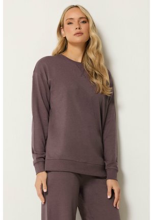 Kvinde med langt blondt hår iført en løs mørkelilla sweatshirt og matchende bukser, stående mod en lys væg.