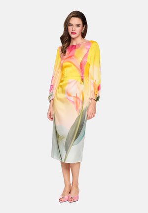 Woman wearing a long-sleeve, midi yellow dress with a pink and green floral print, paired with pink open-toe heels, standing against a white background.