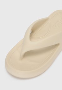 Beige varvasandaalit, joissa on kumimateriaali, V-muotoinen varvastuki, teksturoitu pinta ja sileät, pyöristetyt reunat. Varustettu hillityllä brändilogolla hihnassa.