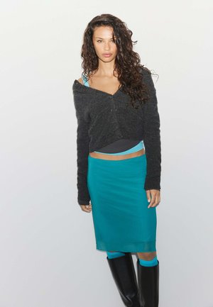 Femme aux longs cheveux bouclés porte un pull gris foncé épaules dénudées, une jupe bleu sarcelle, un haut bleu assorti, et des bottes noires montantes jusqu'aux genoux sur un fond uni.