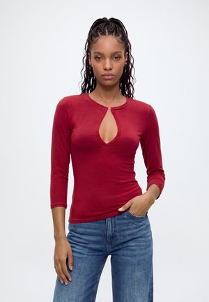 Femme aux cheveux tressés portant un haut rouge ajusté à manches longues avec un col en trou de serrure et un jean bleu, debout devant un fond uni.