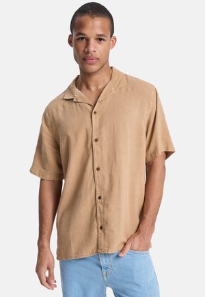 Młody mężczyzna ubrany w jasnobrązową koszulę na krótki rękaw z zapięciem na guziki oraz jasnoniebieskie dżinsy, stoi z jedną ręką w kieszeni na jednolitym tle.