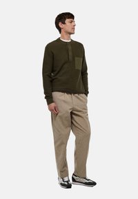 Pull en maille vert olive avec un col rond, trois boutons et une poche poitrine ; associé à un pantalon en velours côtelé beige et des baskets.