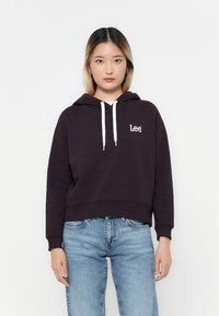Haut à capuche violet raccourci avec un cordon de serrage et un logo blanc sur la poitrine. Porté avec un jean en denim bleu clair, présentant une coupe décontractée.