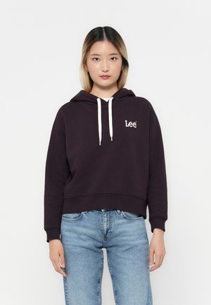 Lilla kort genser med hette og snøring, med hvit logo på brystet. Båret med lyseblå denim jeans med en avslappet passform.