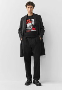 Camiseta gráfica negra con texto rojo debajo de un abrigo gris carbón. Pantalones negros de corte holgado y zapatos negros con cordones completan el atuendo.