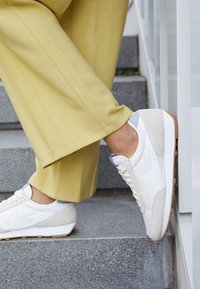Nike-sneakers har en vit och krämfärgad färgkombination med mocka och textilmaterial, en rundad tå och en sula av gummigummi. Matchas med gula byxor.