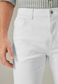 Nahaufnahme einer Person in weißen Jeans und einem grün gestreiften Hemd, mit dem linken Arm entspannt an der Seite.