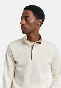 Hellcremefarbener Zip-Hals-Pullover aus weichem Stoff. Merkmale einen Viertel-Reißverschluss und eine strukturierte Oberfläche mit einem dezenten Logo auf der Brust.
