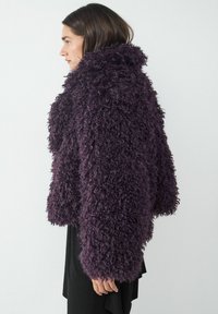 Fioletowy puszysty płaszcz z teksturowanego, kręconego materiału, z szerokim kołnierzem i dużymi rękawami, noszony na czarna bluzkę.