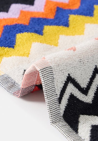 Serviette avec des motifs zigzag colorés en noir, blanc, jaune, bleu, orange et rose, partiellement repliée sur une surface blanche.