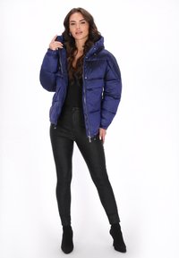 Jachetă matlasată bleumarin cu glugă, fermoar frontal și design matlasat. Asortată cu pantaloni negri conici și botine negre până la gleznă, pe un fundal alb.