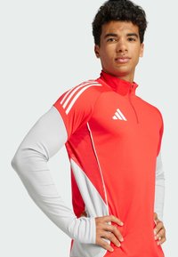 Rød atletisk pullover med halvlynlås, prydet med lange grå ærmer og hvide trekantede detaljer på kroppen. Tre hvide striber på skuldrene.