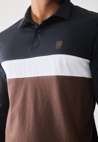 Man wearing a black, white, and brown color-blocked long-sleeve polo shirt with a small emblem on the chest.