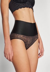 Hög midja svarta underkläder med en slät midjeband och spetsmönstrad nedre del, som visar intrikata blommönster.