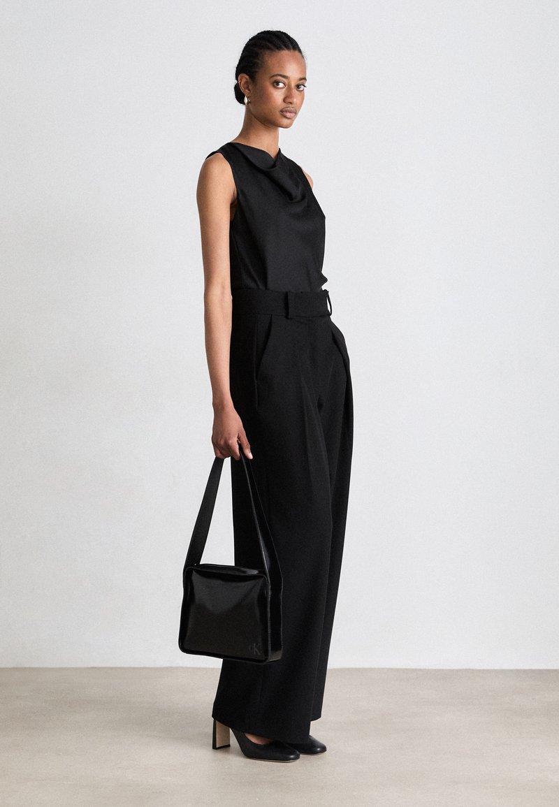 Schwarzes ärmelloses Top mit Drapierung am Hals, kombiniert mit weit geschnittenen schwarzen Hosen und einer glänzenden schwarzen Handtasche. Spitz zulaufende schwarze Absätze vervollständigen den Look.