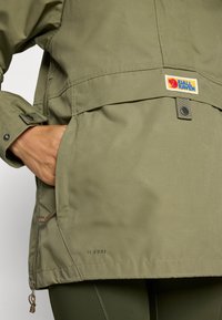 Hand in de zak van een olijfgroen Fjällräven G-1000-jacket, met zichtbare voorflapzak met drukknoop en het merklogo.