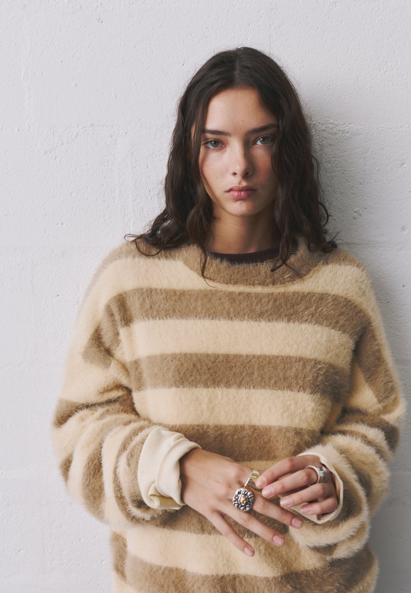 Pehmeä raidallinen neule vaaleassa ja ruskeassa, jossa on ribbikankaiset hihansuujat ja pyöreä pääntie. Mallilla on päällään hopeisia ja kukkakuvioisia sormuksia.