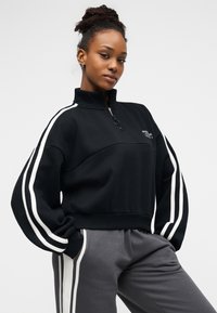 Μαύρο φούτερ με φερμουάρ μέχρι το στήθος, λευκές ρίγες στα μανίκια και κεντημένο λογότυπο στο στήθος. Συνοδεύεται από γκρι παντελόνι φόρμας με αντίστοιχες ρίγες.