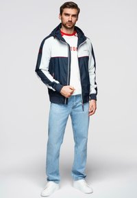Marineblå og hvid hættetrøje med lynlås, røde detaljer, kontrasterende ærmer, båret over en hvid skjorte, kombineret med lyseblå jeans og hvide sneakers.